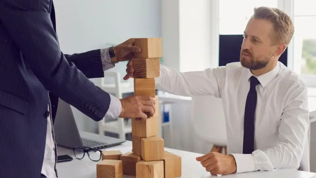 Two professionals in formal wear stacking wooden blocks, symbolizing teamwork and personal growth in a bright office.