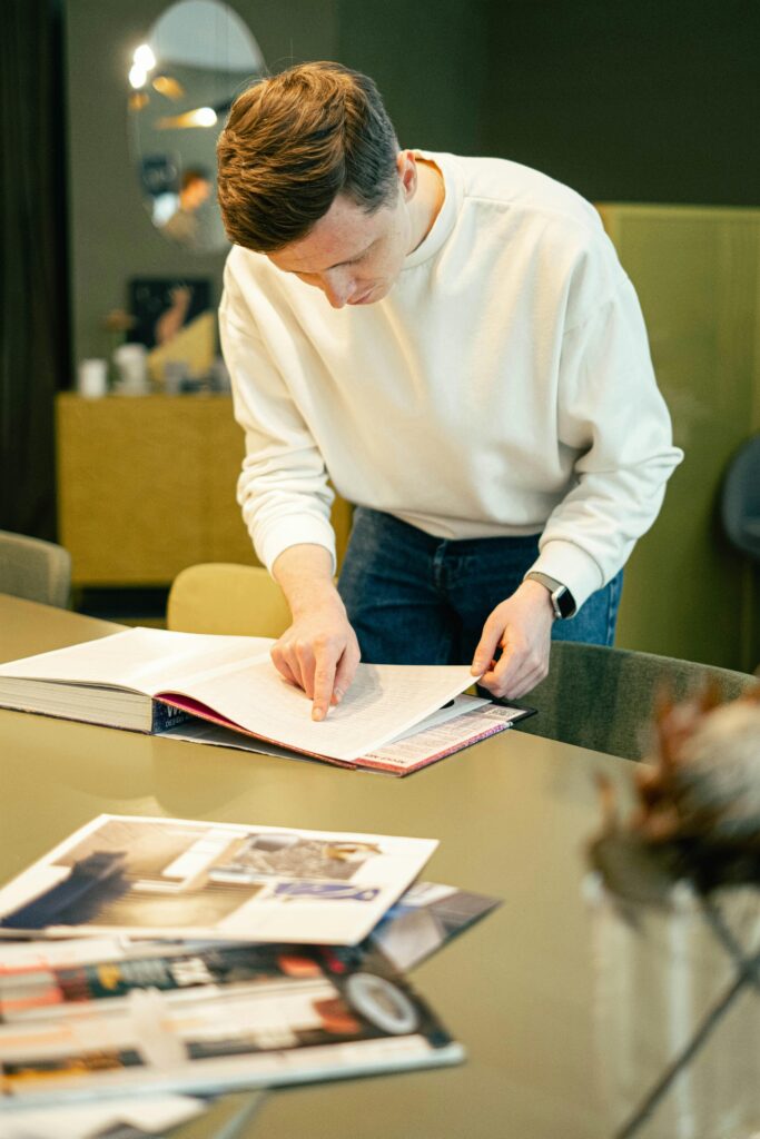 A Man in a white sweater is concentrating on research while reading a book at a creative office desk.